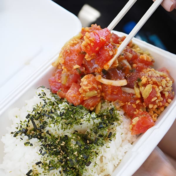 Chopsticks picking up a piece of red poke from a takeout container with rice and furikake.